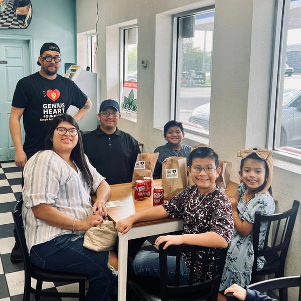 Family posing inside a burger restaurant with branded takeout bags and soft drinks on the table.