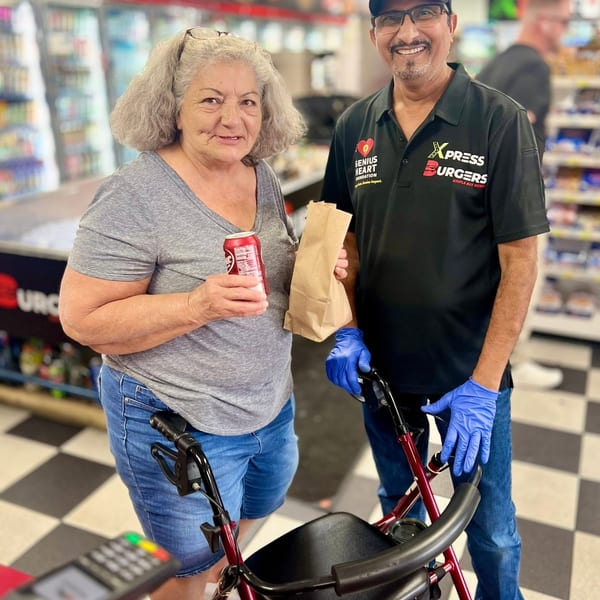 Smiling restaurant worker in gloves stands beside an older woman holding a soda and paper bag inside a grocery store.