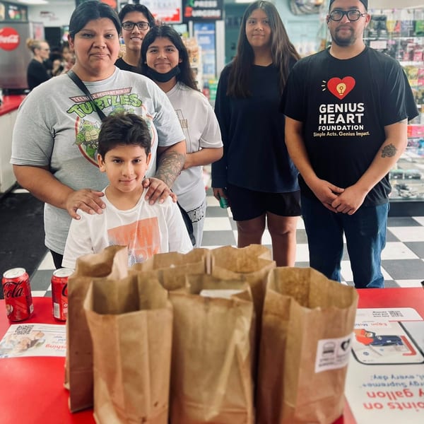 A group of six people poses behind brown paper bags of meals at a casual restaurant.