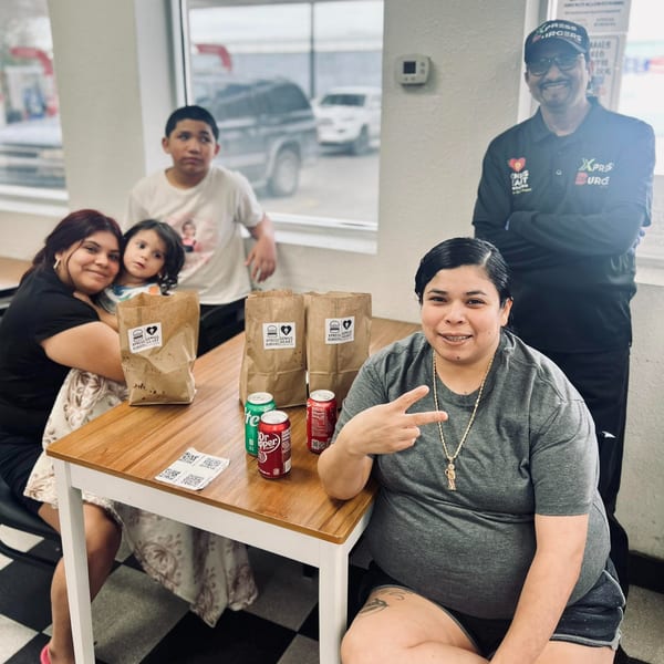 A family with two adults and two children sits at a table with Xpress Genius Burgers bags and sodas.