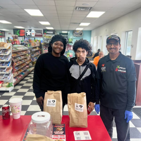 Three Xpress Burgers employees stand behind a counter with takeout bags and drinks in a store.