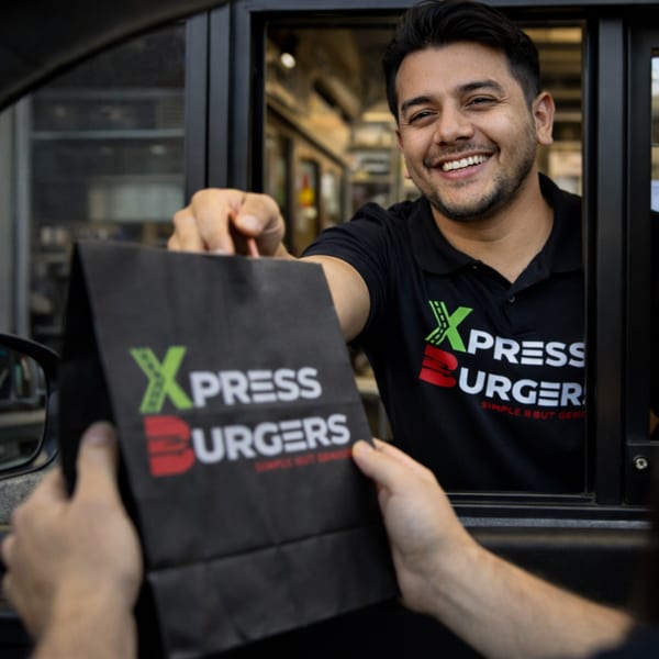 A smiling Xpress Burgers employee hands a customer a branded black bag through a drive-thru window.