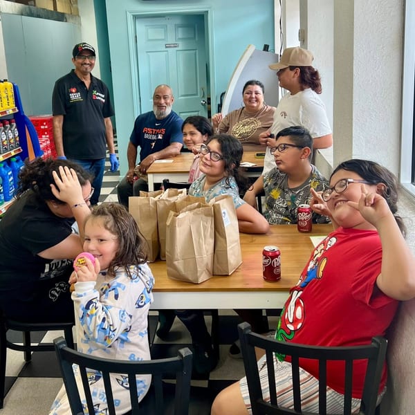 A group of adults and children are seated at tables with brown paper bags and drinks.