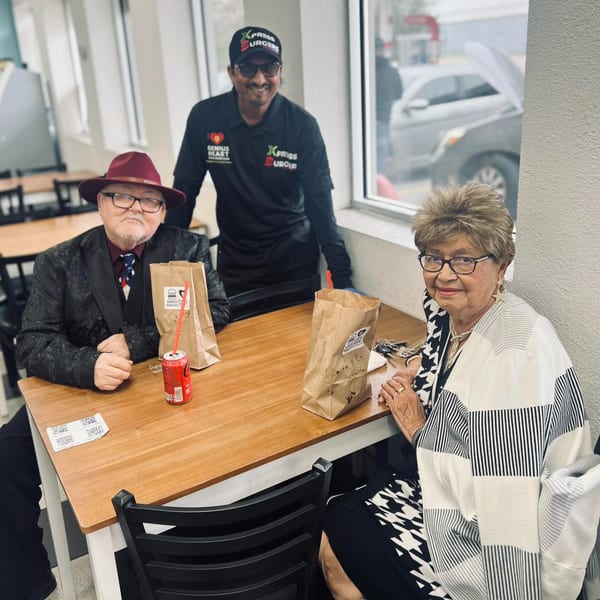 Two customers sit at a table with food bags, while a smiling Xpress Burgers employee stands behind them.