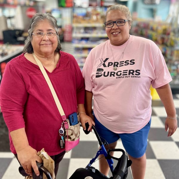 Two women, one in a pink Xpress Burgers shirt, smiling in a store with checkered floors.