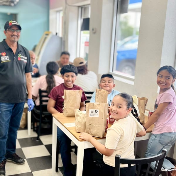 A man in a black polo shirt and cap stands smiling next to children seated at tables with food bags.